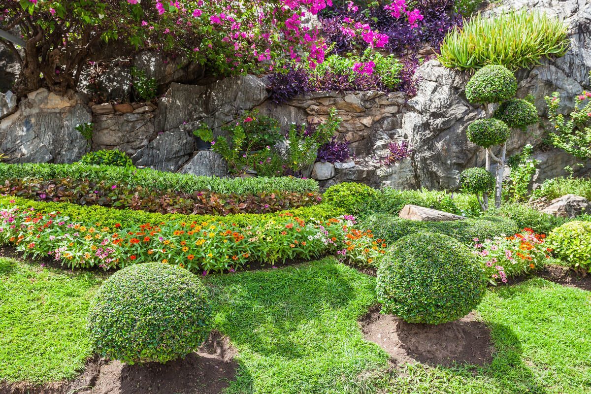 Un jardín vibrante con vegetación en capas, flores coloridas y arbustos con formas sobre un fondo rocoso.