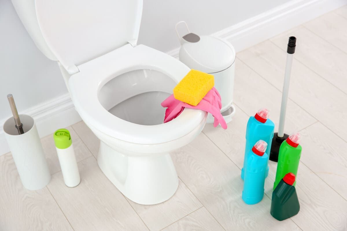 Un inodoro que se limpia con diversos suministros: cepillo de baño, botellas de spray, esponja y guantes.