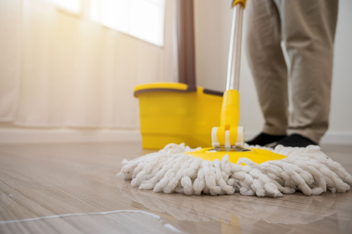 Persona fregando un piso de madera con un trapeador amarillo y un balde en una habitación con luz natural.