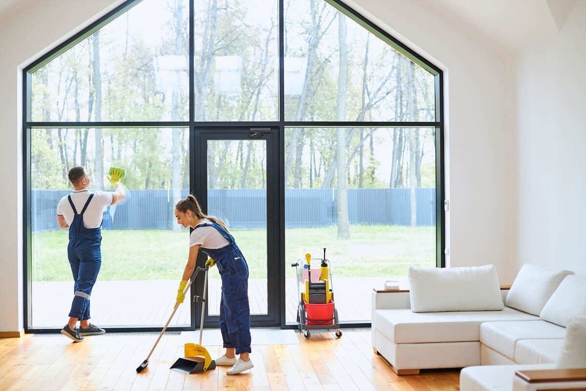 Dos personas limpiando una habitación muy iluminada con grandes ventanas: una lavando una ventana y la otra barriendo el suelo.