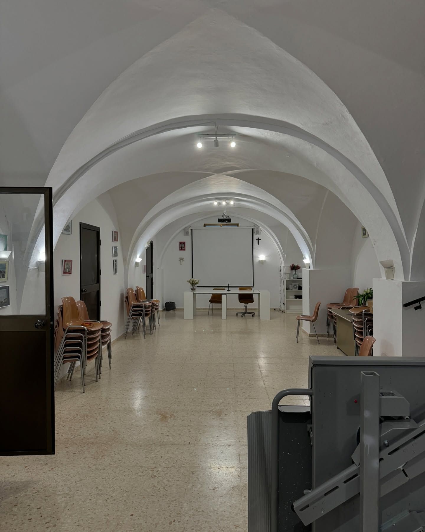 Sala blanca larga y arqueada con sillas, mesas y pantalla de proyección.