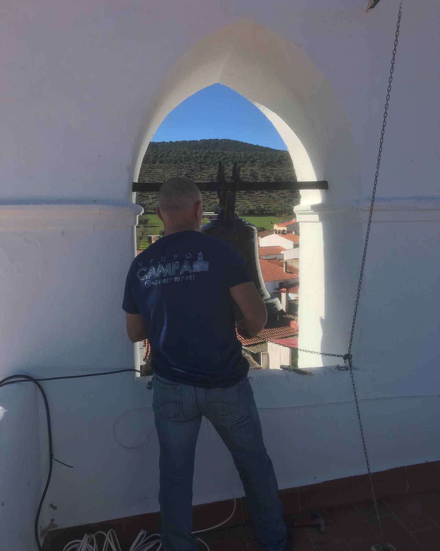 Hombre dentro de una torre blanca, mirando desde una ventana arqueada hacia un pueblo y una ladera.