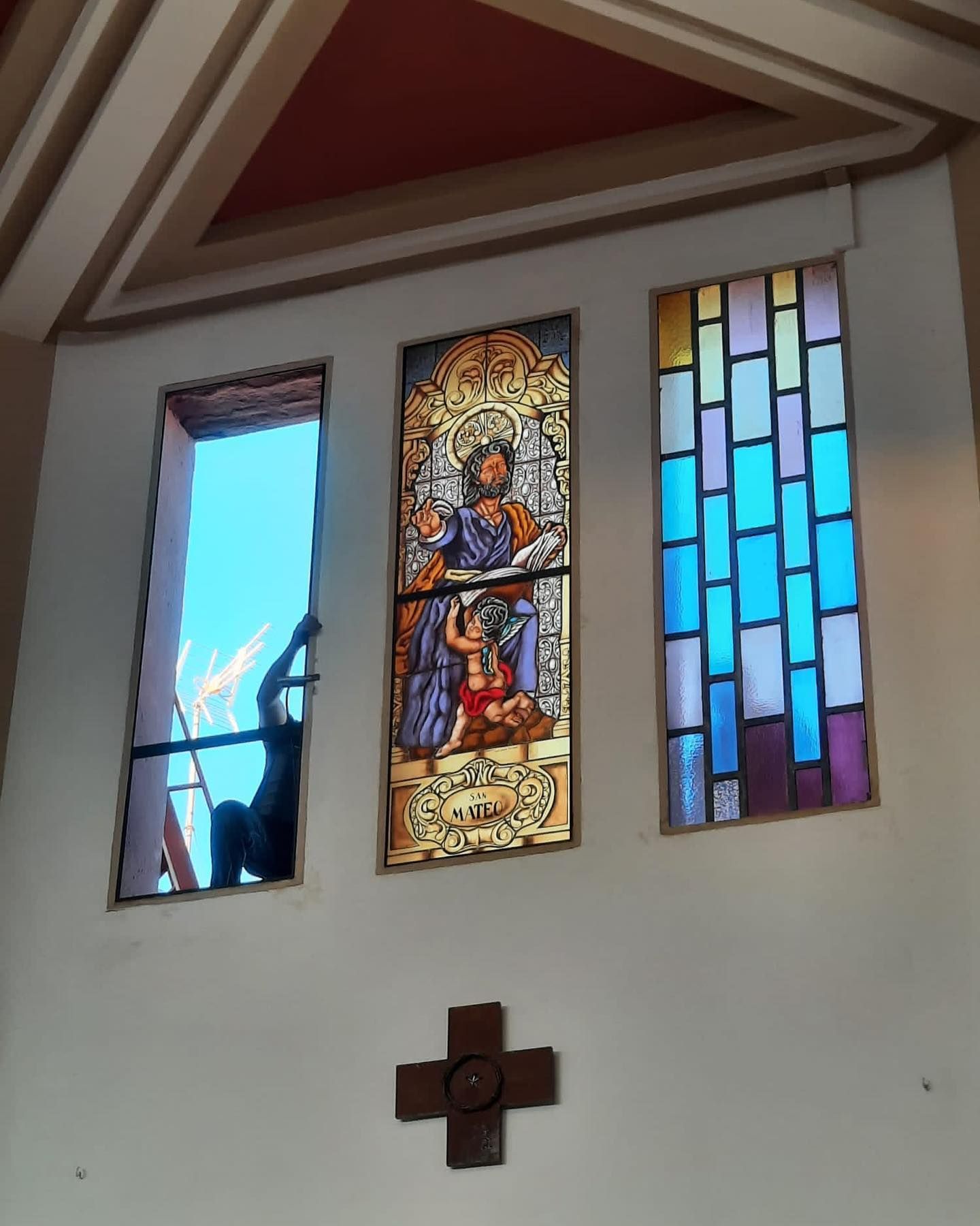 Tres vitrales en una iglesia. Una persona trabaja en el vitral izquierdo, abierto. Debajo hay una cruz.