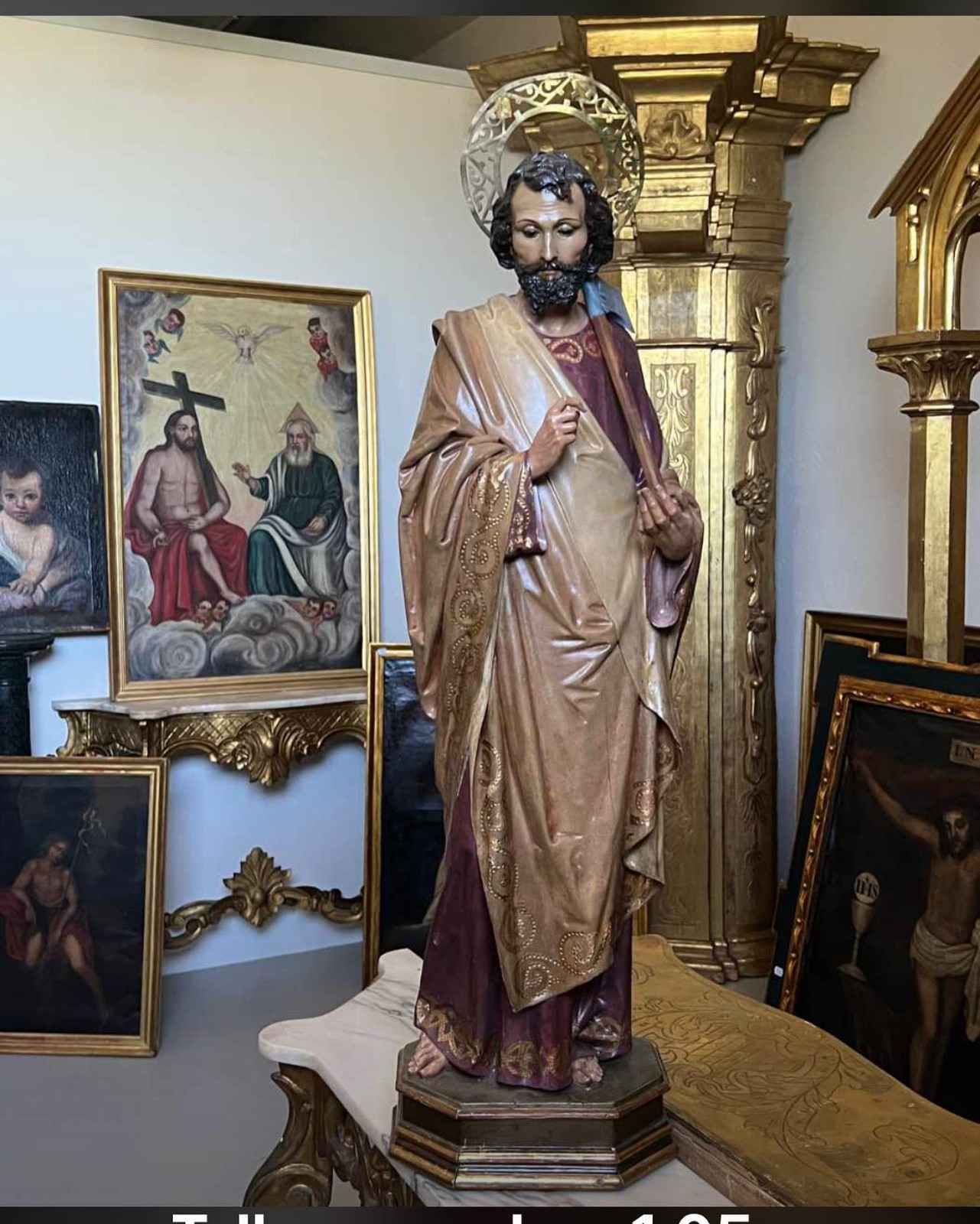 Estatua de un santo con aureola, sosteniendo una llave. Vestida con túnicas marrones, de pie en una sala con pinturas y detalles dorados.