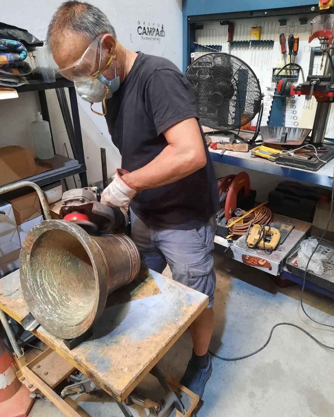 Un hombre con equipo de seguridad lija una campana de bronce en un taller.
