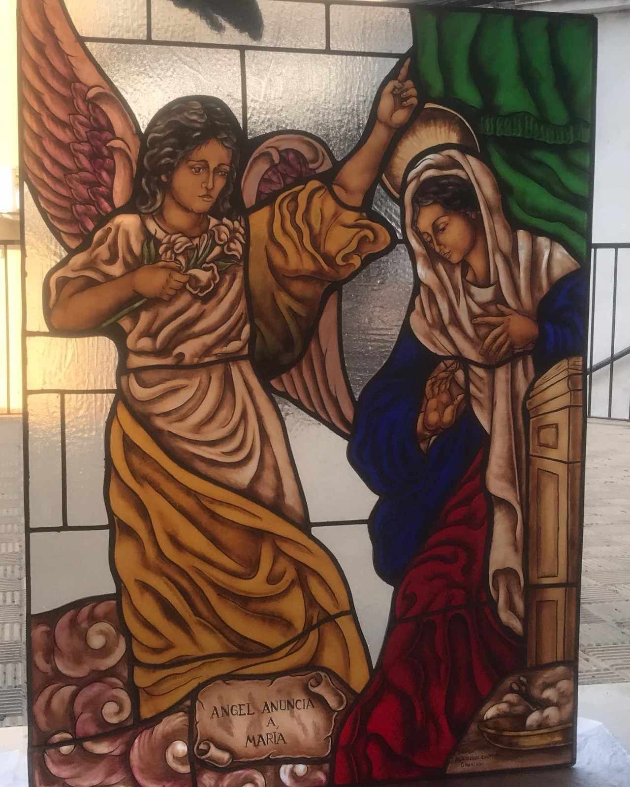 Escena de vidriera: ángel con la mano levantada hace gestos hacia María, vestida de azul y blanco, en un arco.