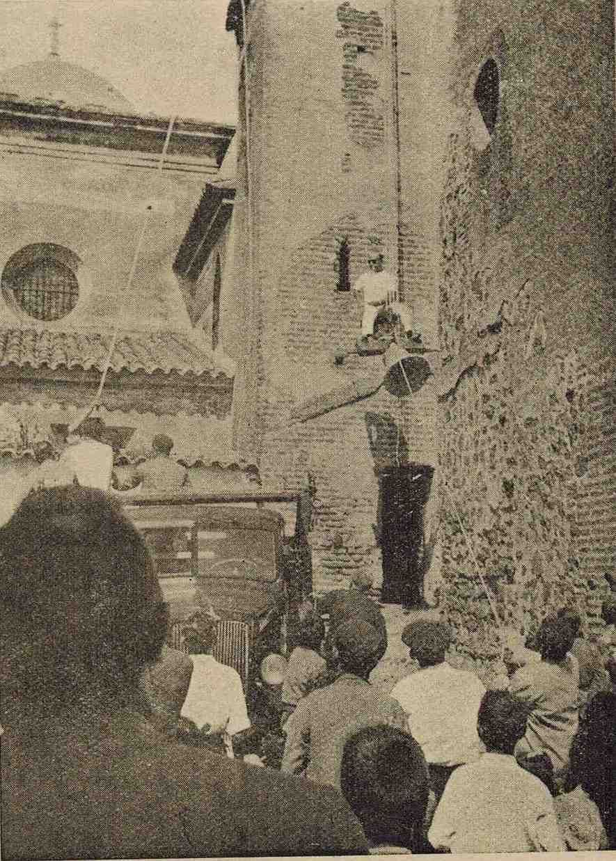 Una multitud se reúne y mira hacia arriba, hacia una alta torre de ladrillos que hay al lado de un edificio con techo abovedado.