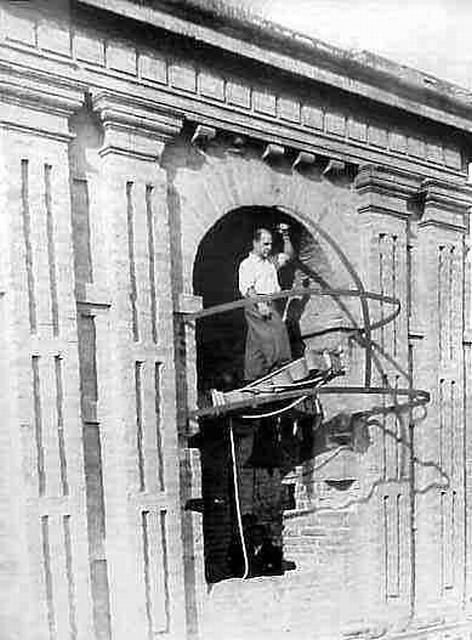 Hombre trabajando en una estructura de metal en espiral afuera de la ventana arqueada de un edificio de ladrillo.
