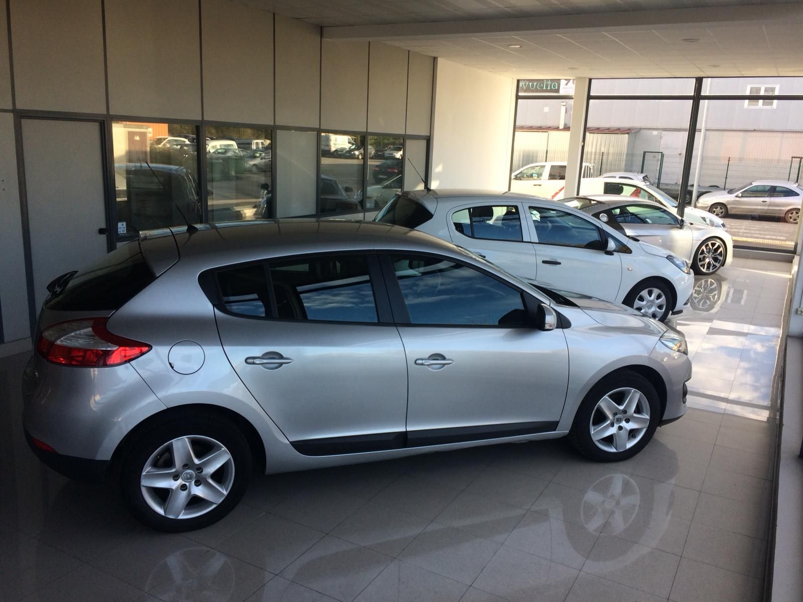Un automóvil hatchback plateado en una sala de exposición, con otros dos automóviles visibles al fondo.