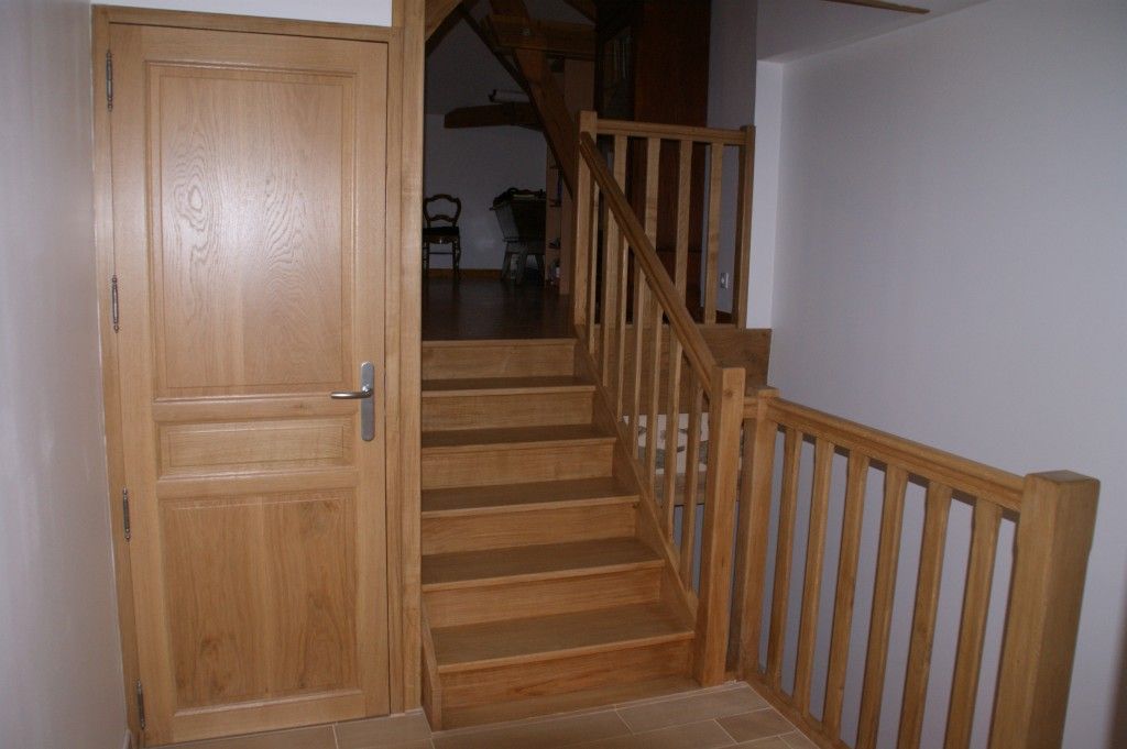 Une porte en bois se trouve à côté d'un escalier en bois avec une rampe assortie menant à une pièce à l'étage faiblement éclairée.