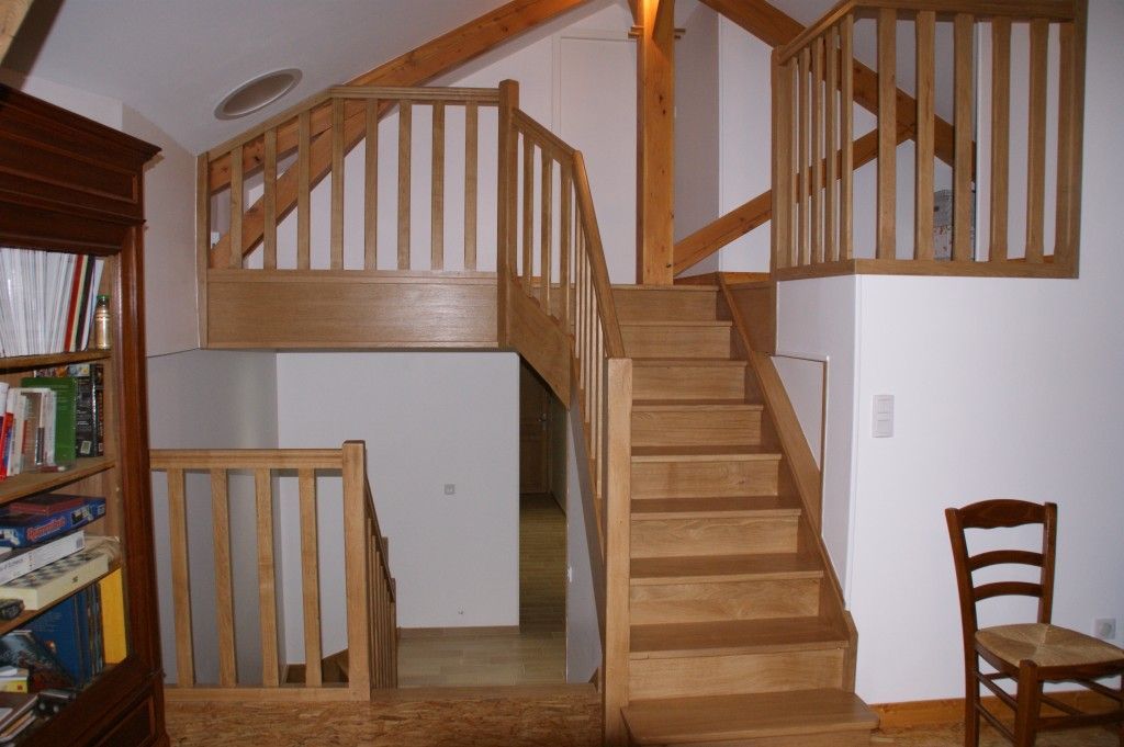 Un escalier en bois menant à une mezzanine, avec une rampe assortie et une bibliothèque, dans un intérieur lumineux et moderne.