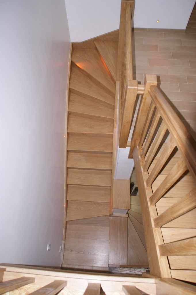 Vue plongeante d'un escalier en bois clair avec une rampe en bois, descendant dans un couloir lumineux aux tons neutres.