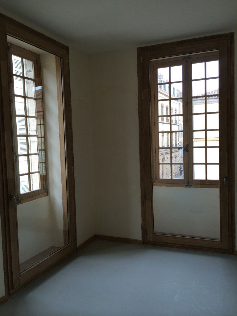 Un coin de pièce aux murs clairs et au sol gris, avec deux hautes fenêtres à cadres en bois.