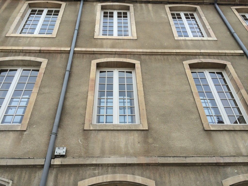 La façade beige du bâtiment présente deux rangées de fenêtres cintrées à plusieurs carreaux avec des cadres blancs et une gouttière verticale grise.