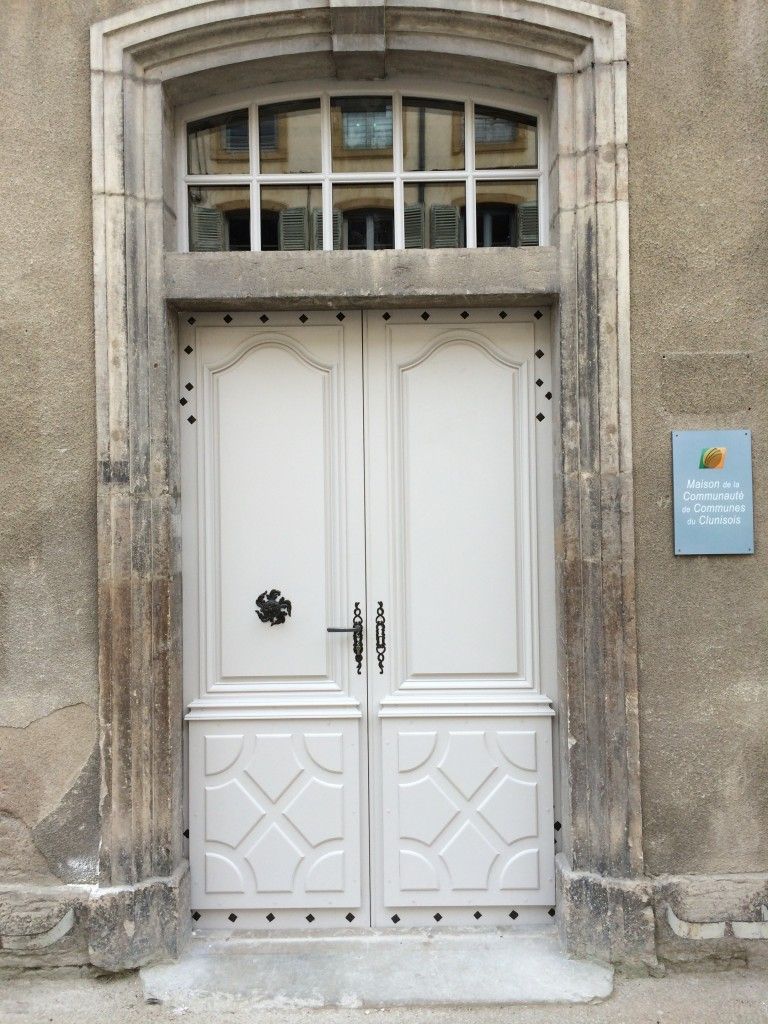 Une double porte gris pâle encastrée dans une arche en pierre, avec une imposte et une petite enseigne sur le mur adjacent.