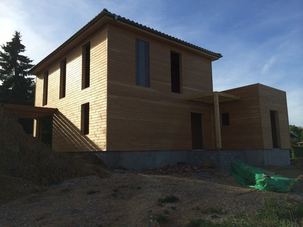 Une maison à deux étages, avec un bardage en bois clair et des boiseries foncées, est en construction sur un terrain vague sous un ciel d'un bleu éclatant.