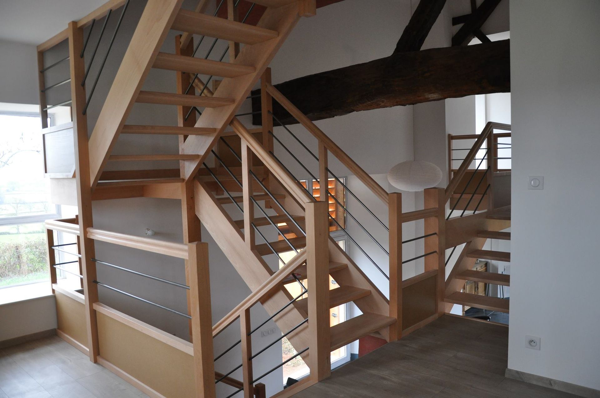 Un escalier en bois clair avec des rampes modernes en câbles métalliques dans un intérieur lumineux et ouvert avec une poutre apparente au plafond.