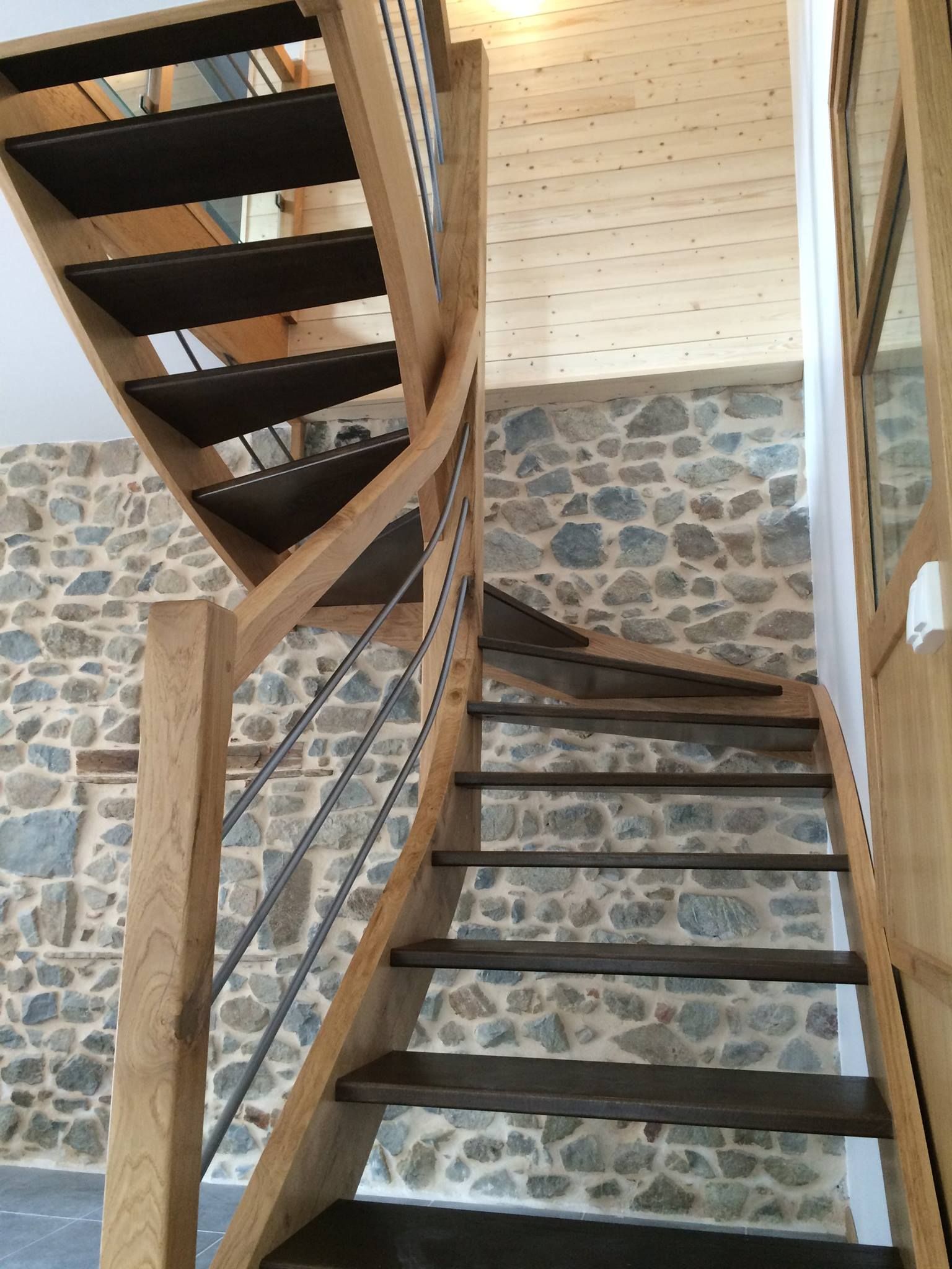 Escalier en bois avec marches en métal foncé et rampes en câbles, adossé à un mur en pierre texturée à l'intérieur d'une maison.
