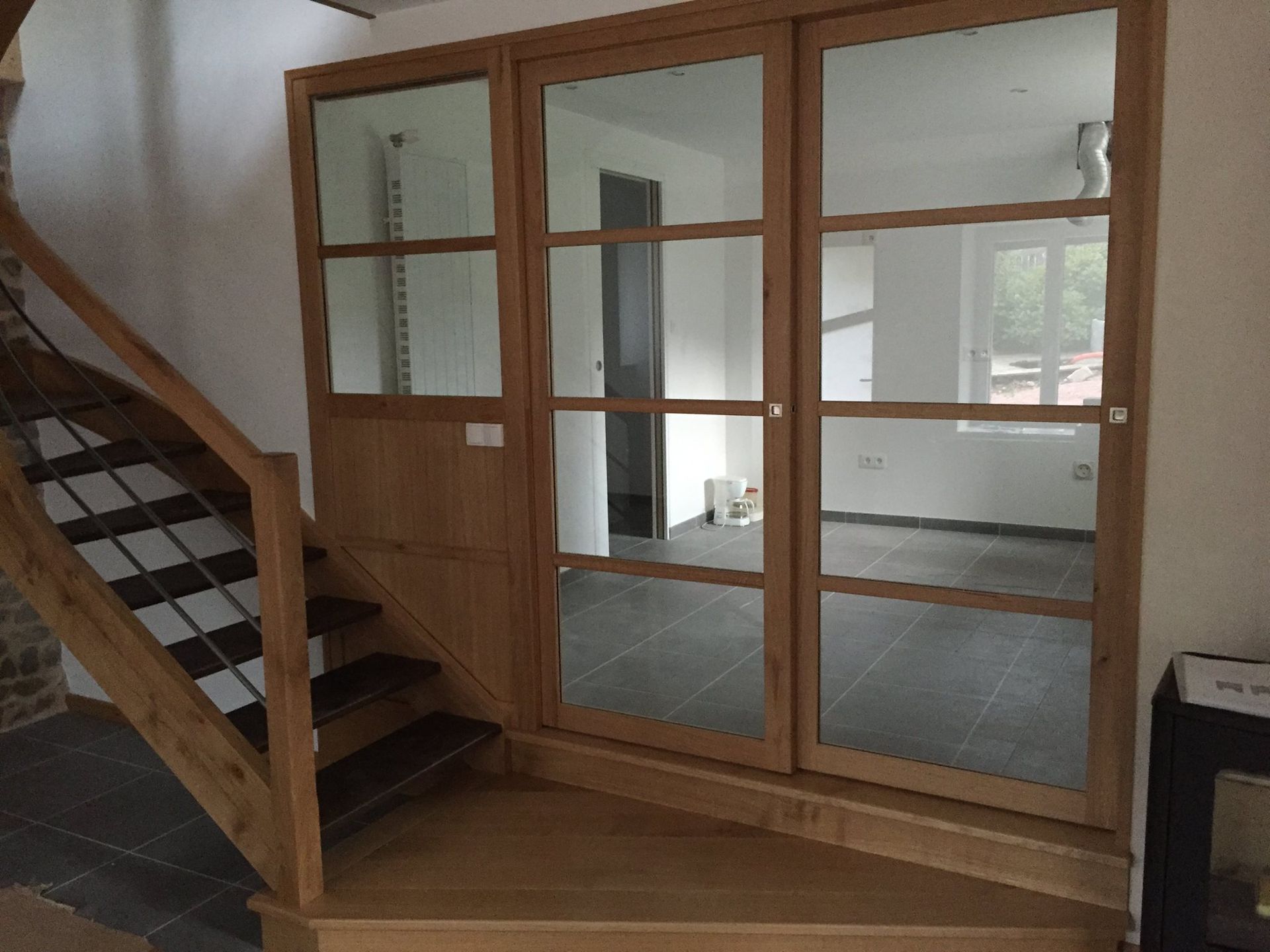 Un escalier en bois jouxte une cloison vitrée à cadre en bois, dans une pièce au sol carrelé.