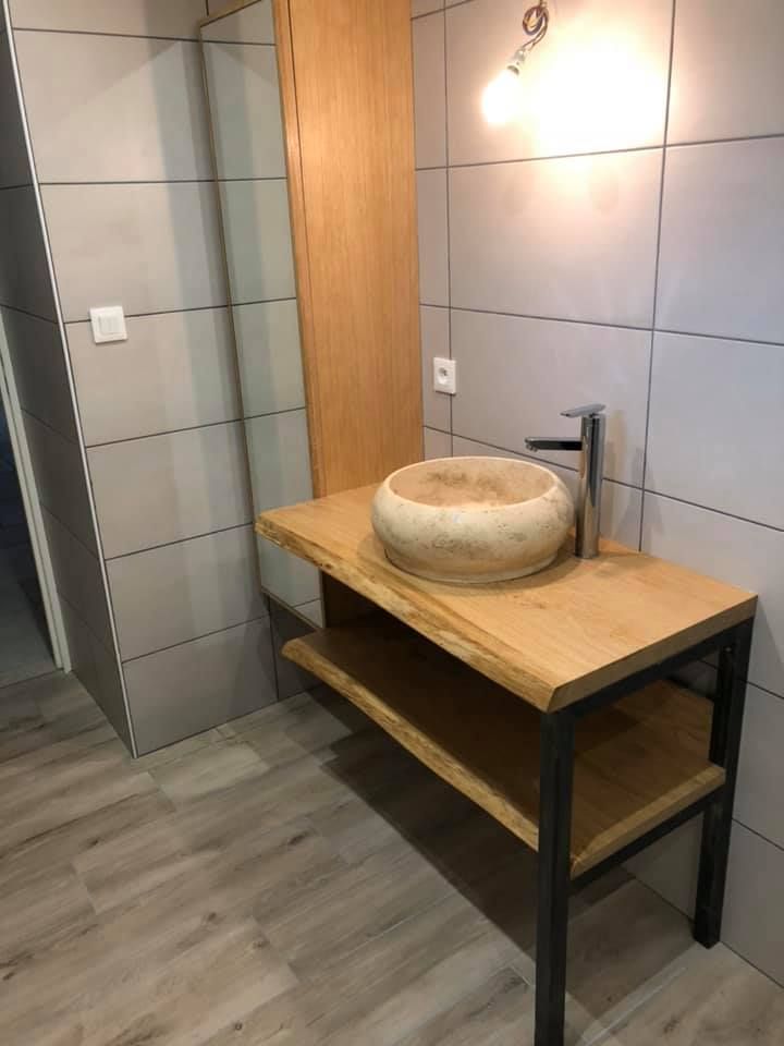 Un lavabo rustique en pierre est posé sur un meuble-lavabo en bois aux pieds en métal noir, devant un mur de salle de bains carrelé de gris.