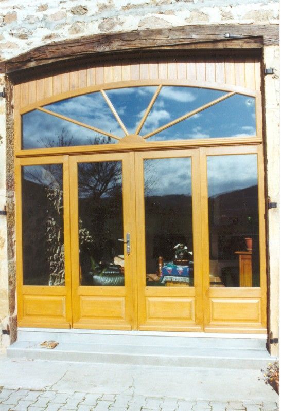 Un ensemble de portes doubles en bois brun clair avec panneaux de verre et une imposte cintrée en forme de soleil, encastrées dans un mur en pierre.