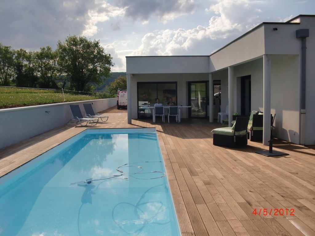 Maison moderne avec piscine, terrasse en bois, mobilier de jardin et pelouse sous un ciel nuageux.