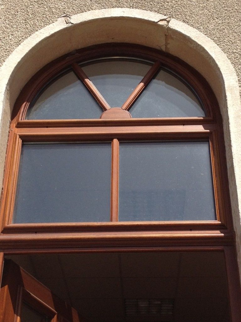 Une fenêtre cintrée en bois brun, avec un meneau central et une partie supérieure en forme d'éventail, encastrée dans un mur en pierre beige texturée.