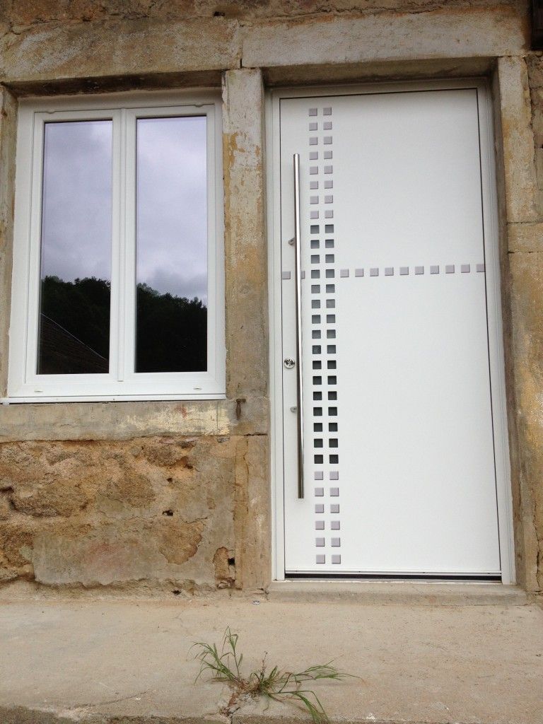 Une porte moderne blanche ornée d'une grille verticale de motifs carrés et d'une longue poignée, placée à côté d'une fenêtre dans un mur en pierre.