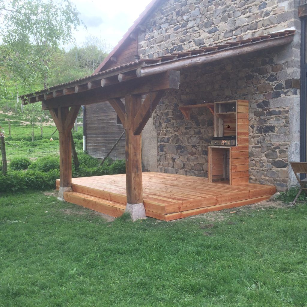 Une terrasse en bois avec un porche couvert attenant à un bâtiment en pierre, comprenant un petit meuble de cuisine extérieure en bois.