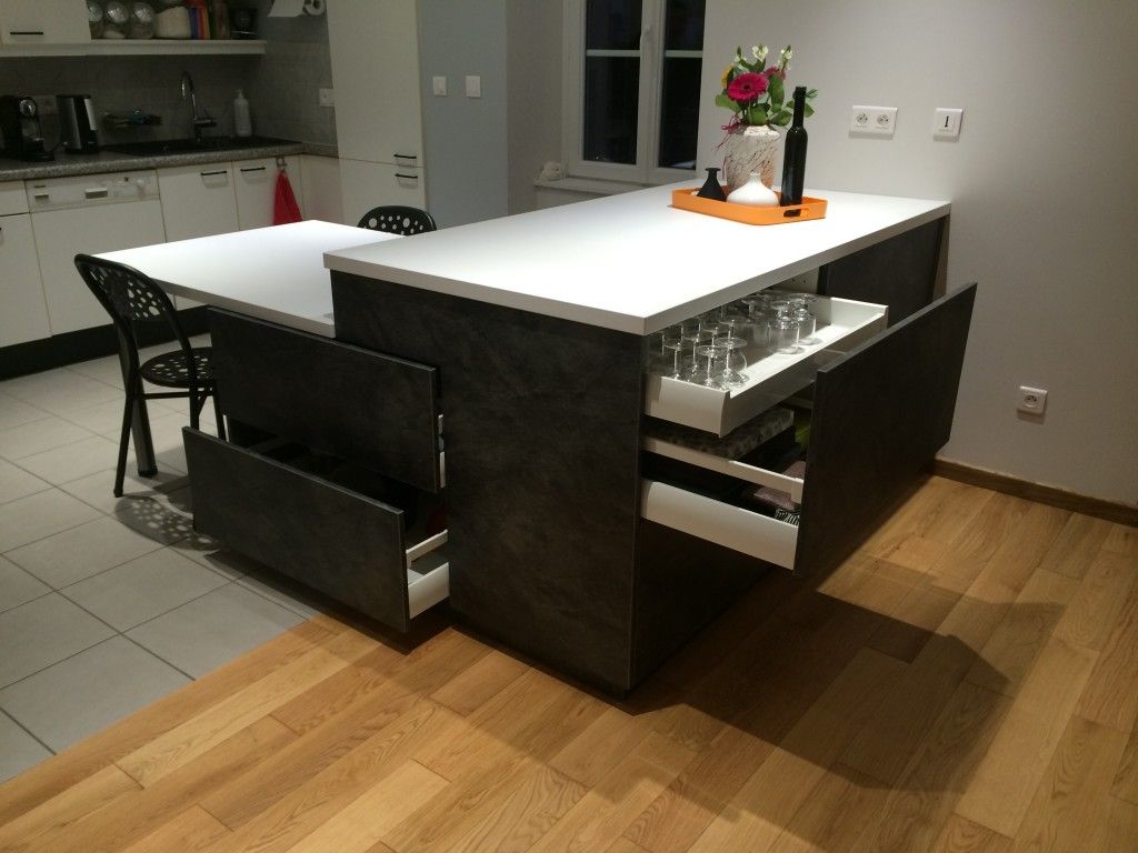 Un comptoir de boulangerie en forme de L avec des vitrines en verre, des plans de travail en bois et des panneaux gris devant un mur garni d'étagères.