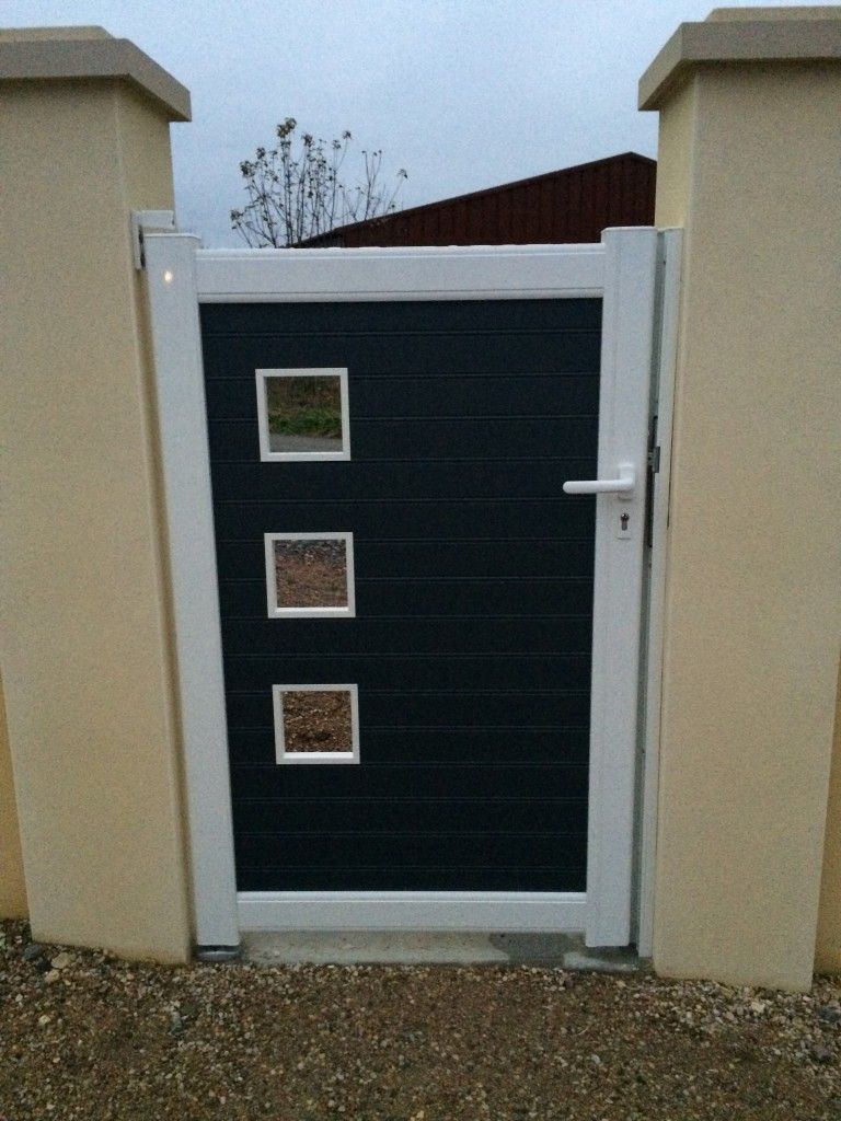 Un portillon piéton gris foncé avec trois fenêtres carrées dans un cadre blanc, installé entre deux piliers clairs.