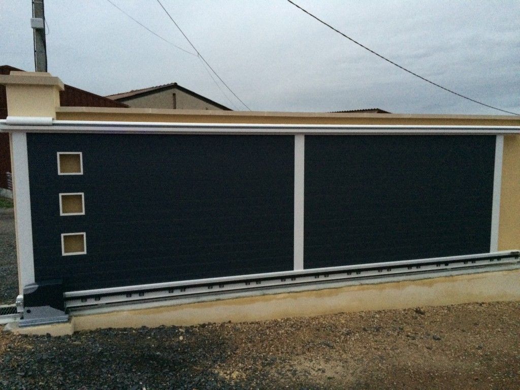 Un portail coulissant pour allée, avec des lattes horizontales gris foncé et trois petites fenêtres carrées sur la gauche, à côté d'un mur beige.