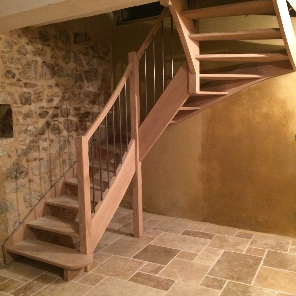 Escalier en bois en forme de L avec balustres métalliques, adossé à un mur en pierre et à un mur lisse couleur beige sur un sol carrelé en pierre.