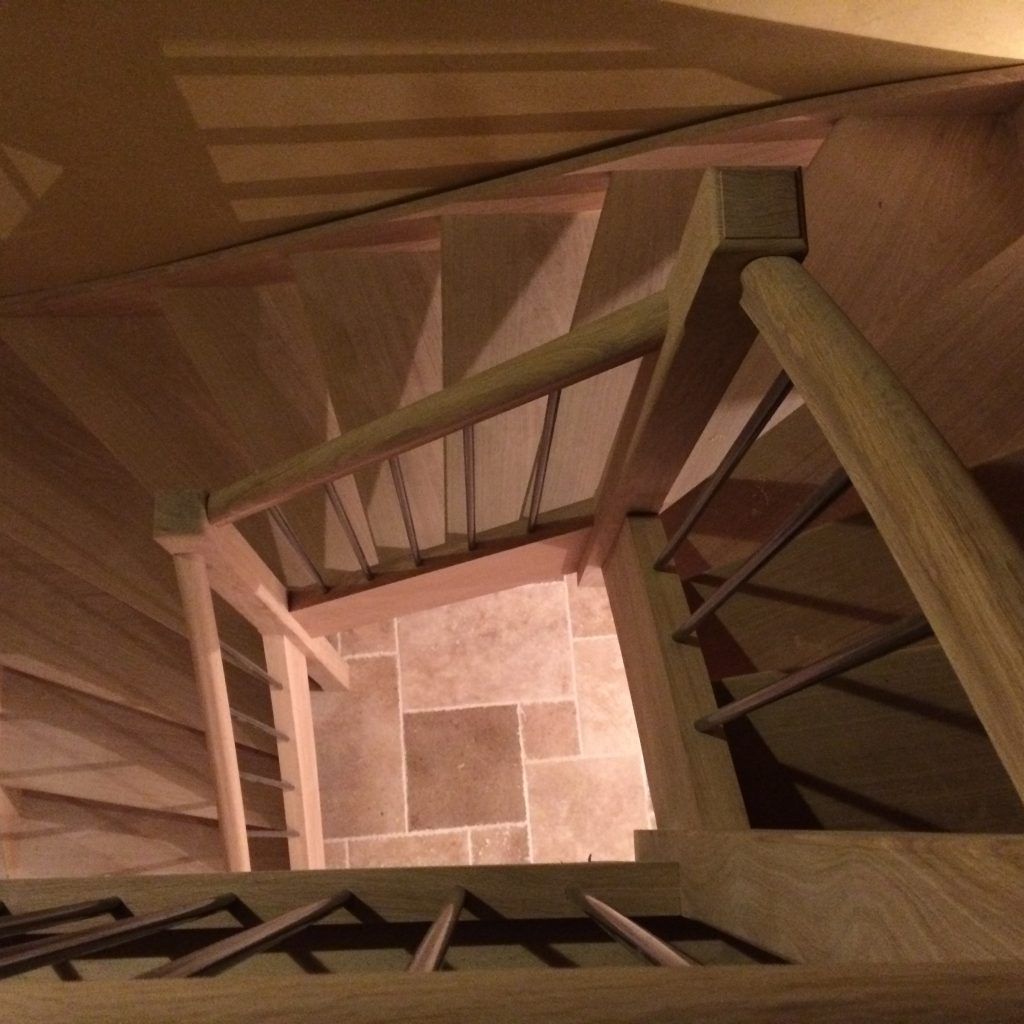 Vue en plongée d'un escalier en colimaçon en bois avec des rampes métalliques donnant sur un palier carrelé carré.