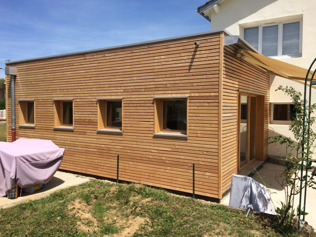 Une extension de maison moderne, revêtue de bois, avec quatre fenêtres carrées et une grande porte coulissante en verre, attenante à une maison blanche.