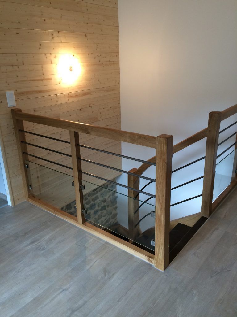 Un palier d'escalier doté d'une rampe en bois avec des barres métalliques horizontales et des panneaux de verre, se détachant sur des murs en bois clair.