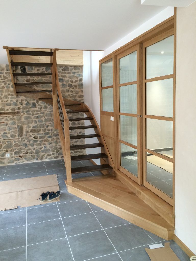 Un escalier moderne en bois aux marches sombres adossé à un mur en pierre, jouxte une grande cloison vitrée à cadre en chêne clair.