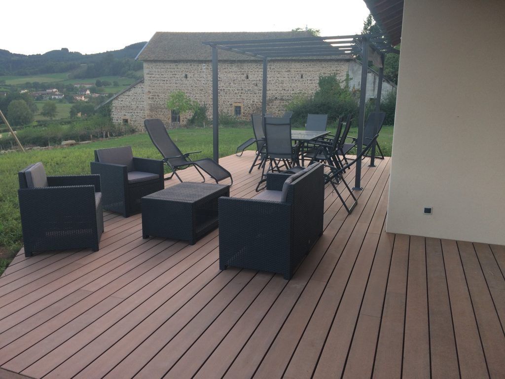 Un patio moderne avec du mobilier sombre et une terrasse en bois, agrémenté d'une pergola et offrant une vue pittoresque sur un bâtiment en pierre et des collines.