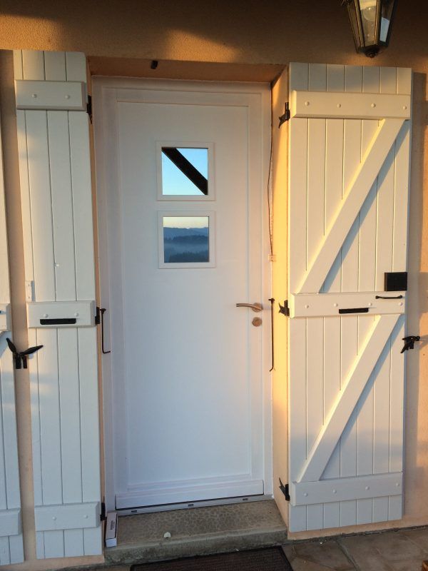 Une porte d'entrée blanche avec deux fenêtres carrées, flanquée de volets en bois blanc fermés, sur un mur extérieur aux tons chauds.