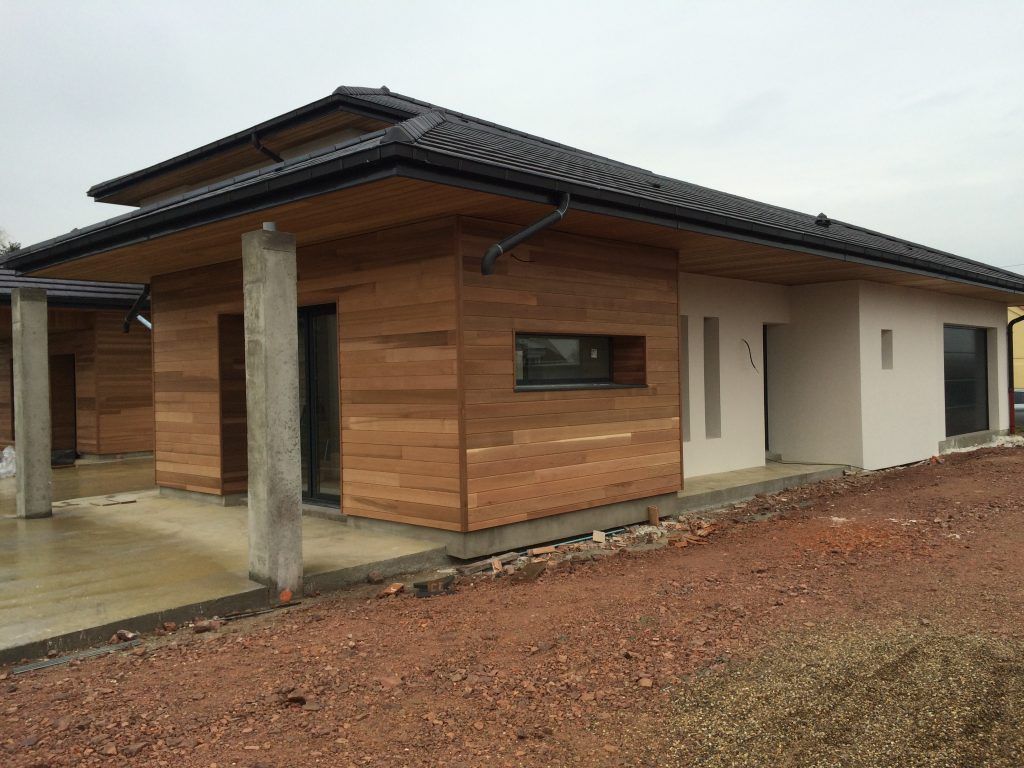Une maison moderne de plain-pied avec un bardage en bois horizontal, un toit à deux pans sombre, des piliers en béton et une cour en terre battue.