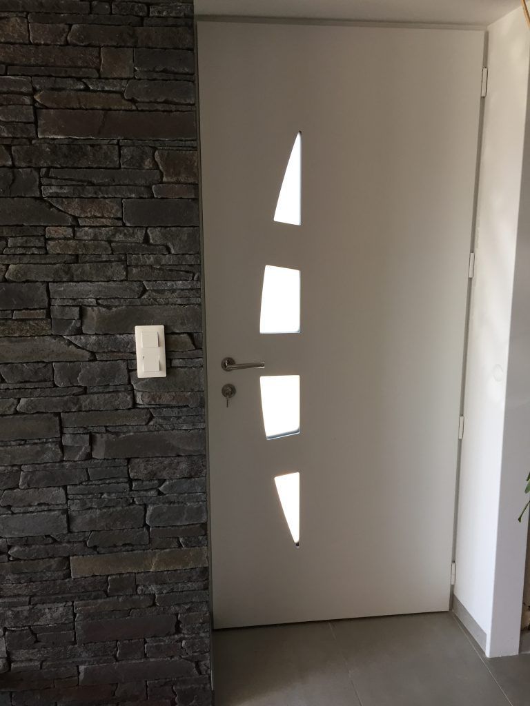 Une porte intérieure blanche percée de quatre fenêtres verticales géométriques, placée à côté d'un mur en pierre empilée gris foncé.