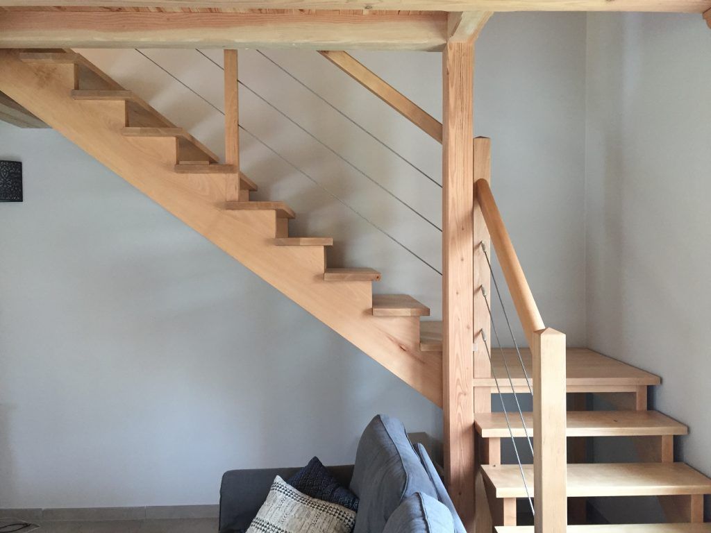 Escalier en bois clair avec rampe à câbles, présentant une conception à contremarches ouvertes, au-dessus d'un canapé gris dans un intérieur de maison.