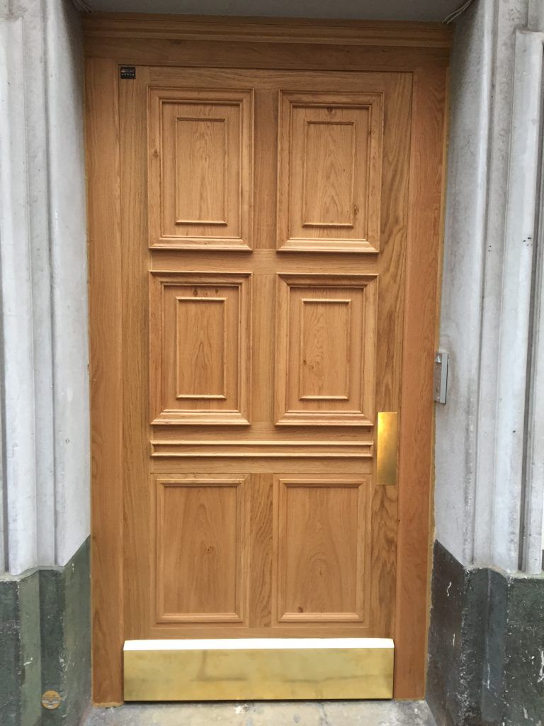 Une porte en bois clair à six panneaux rectangulaires, avec une plaque de protection en laiton et une poignée de porte en laiton.