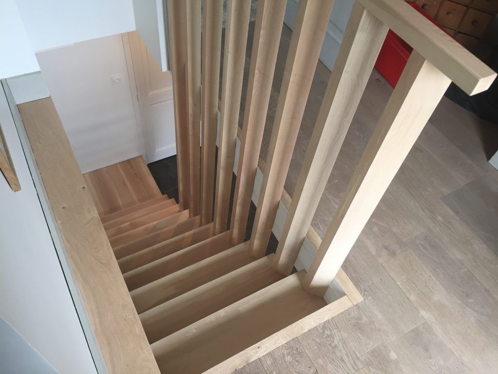 Vue en plongée d'un escalier en bois à rampes verticales descendant vers un étage inférieur dans une maison baignée de lumière.