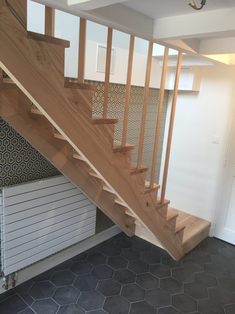 Escalier en bois clair avec de fines rampes verticales, le long d'un mur à motifs, et radiateur blanc sur un sol en carrelage hexagonal noir.