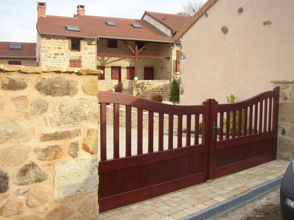 Un portail en bois brun foncé se dresse devant une maison rustique en pierre au toit de tuiles.
