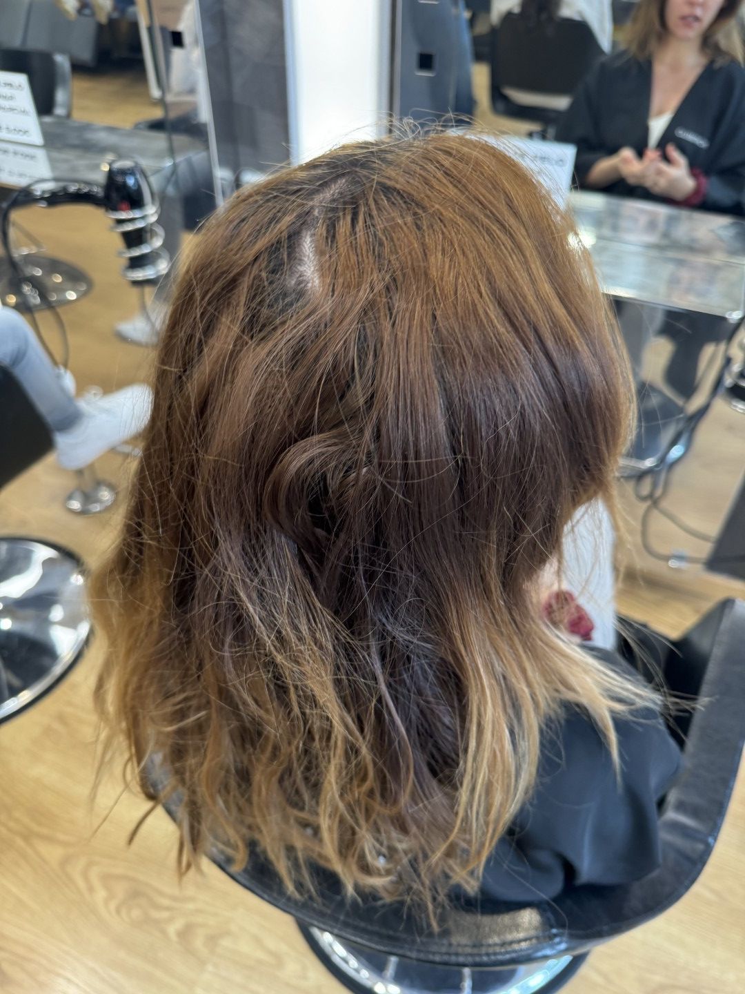 Persona con cabello castaño ondulado sentada en una silla de peluquería, vista desde atrás en un salón de belleza.