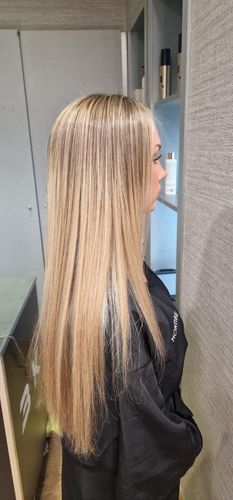 Persona con cabello rubio largo de pie en un pasillo, frente a una pantalla o dispositivo montado en la pared.