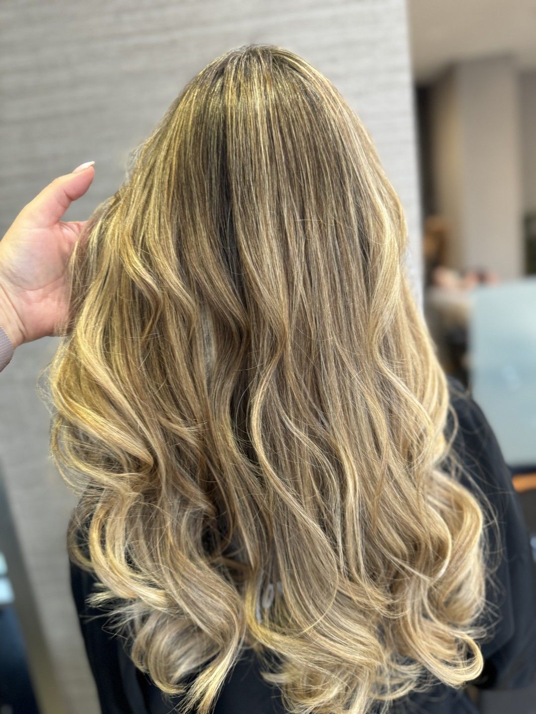 Cabello rubio largo y ondulado visto desde atrás, sostenido por una mano en un salón de belleza.