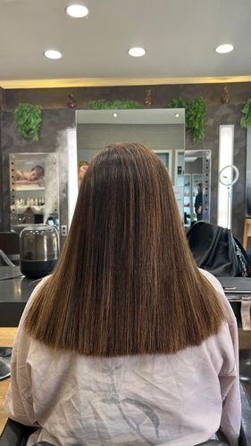 Persona con cabello largo y rizado sentada en una silla de peluquería, vista desde atrás, con espejos y luces al frente.